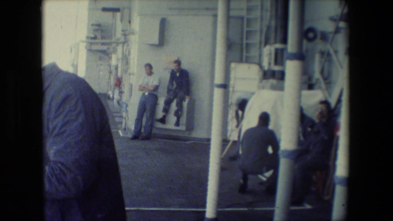 1981 8mm footage frame showing sailors on a naval vessel deck, with railings, ship fittings, vertical poles, and a partially visible figure in the foreground.