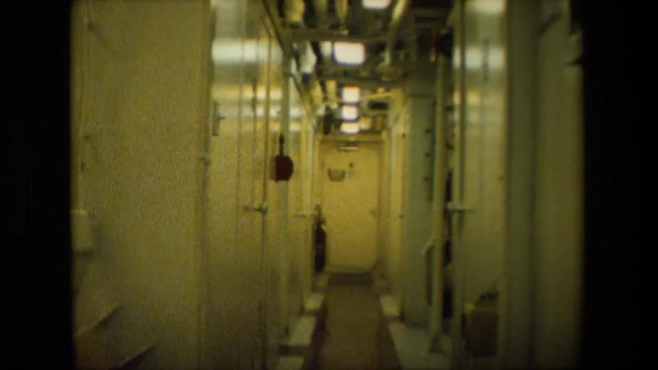 Below-deck ship corridor in yellowish 8mm color, showing metal doors, overhead lighting, wall-mounted fixtures, and pipes along the passageway.