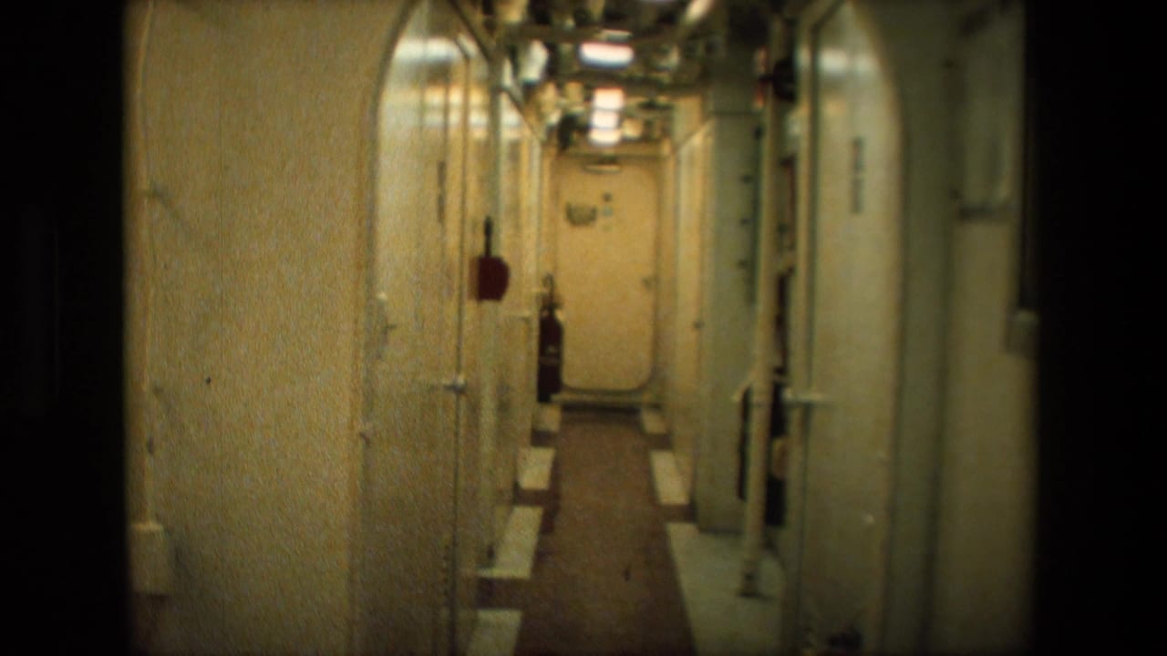 Narrow dimly lit metal corridor in archival 8mm footage, with rounded doorways, exposed piping, wall fixtures, and fluorescent ceiling lights.