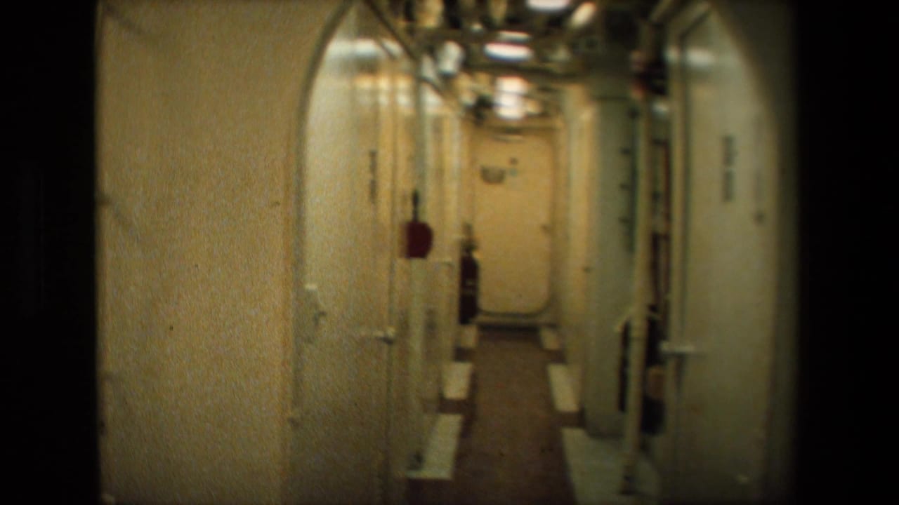 Archival 8mm view down a confined metal passageway, showing overhead piping, fluorescent light, side doors, and a closed door at the far end.