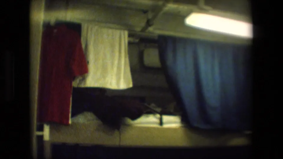 Cramped crew-quarters-style bunk area in archival 8mm footage, with a blue curtain, red shirt, white towel, fluorescent light, and bedding visible.