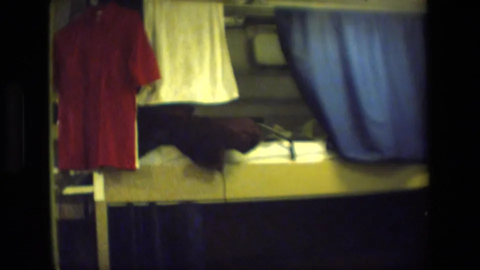 Archival 8mm frame of a compact bunk area, showing a hanging red shirt, white towel, blue privacy curtain, dark bedding, and metal interior surfaces.