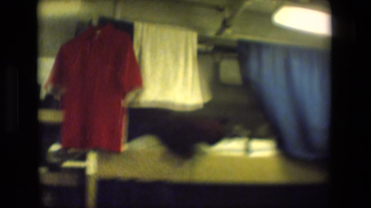Soft 8mm frame of a cramped dormitory or barracks-style room with a red shirt, white towel, bunk bedding, and a blue curtain divider.