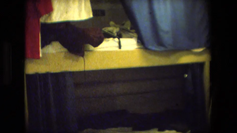 Close handheld frame showing the lower portion of a small sleeping area, with a red garment, white towel, bedding, and blue curtain visible.