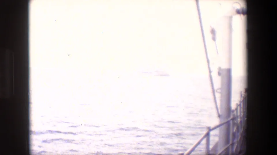 Bright, washed-out ocean view filmed from a vessel, with deck rail, vertical rigging or pipework, and a faint distant shape near the horizon.