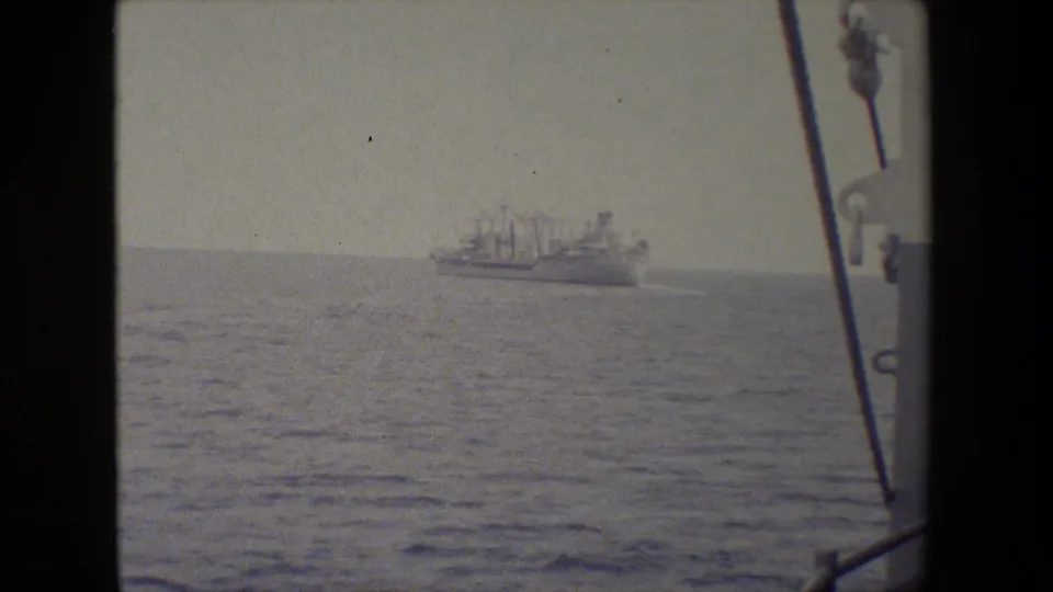 A distant vessel sits on the horizon across open water, framed by the rail or rigging of the filming ship in soft 8mm exposure.