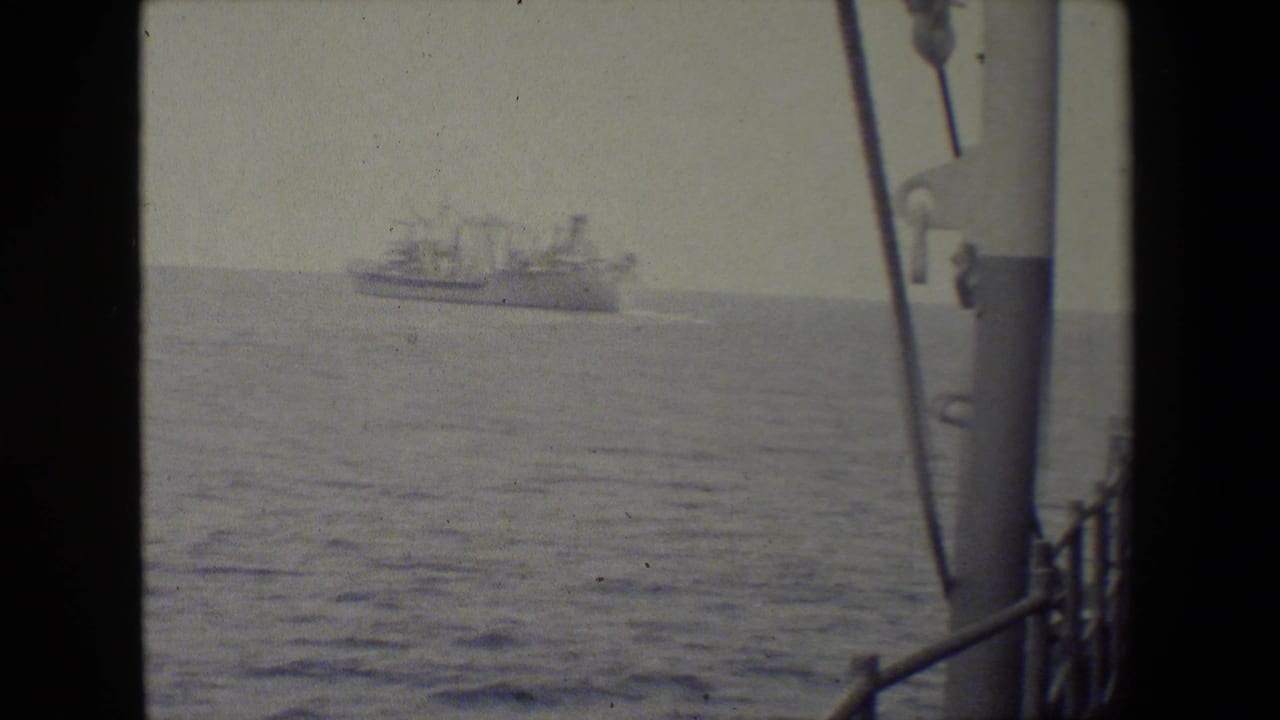 Soft 8mm frame of a distant vessel crossing calm water, filmed from a ship deck with railings and rigging visible along the right edge.