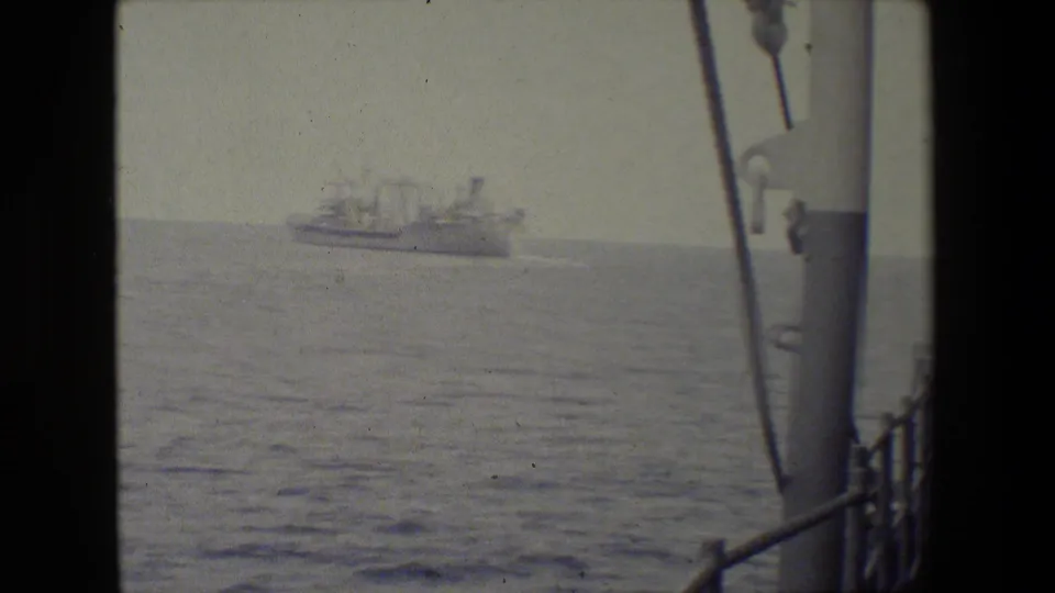 Soft 8mm frame of a distant vessel crossing calm water, filmed from a ship deck with railings and rigging visible along the right edge.