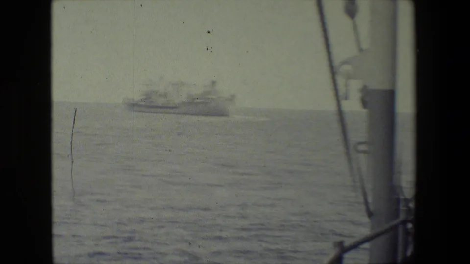 Distant vessel appears on a hazy horizon across calm water, with foreground rigging and deck structure framing the shipboard viewpoint.
