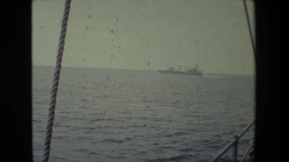 Wide shipboard view over calm open water, with a foreground rope and railings framing a small distant vessel on the horizon.