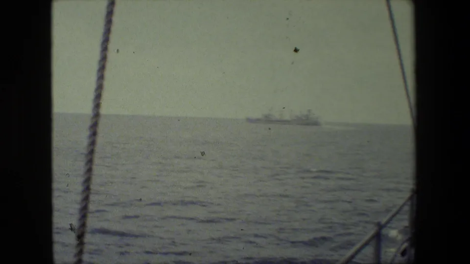 Shipboard home-movie frame looking across calm water toward a distant vessel, with rigging, railing, and dark scan borders visible.