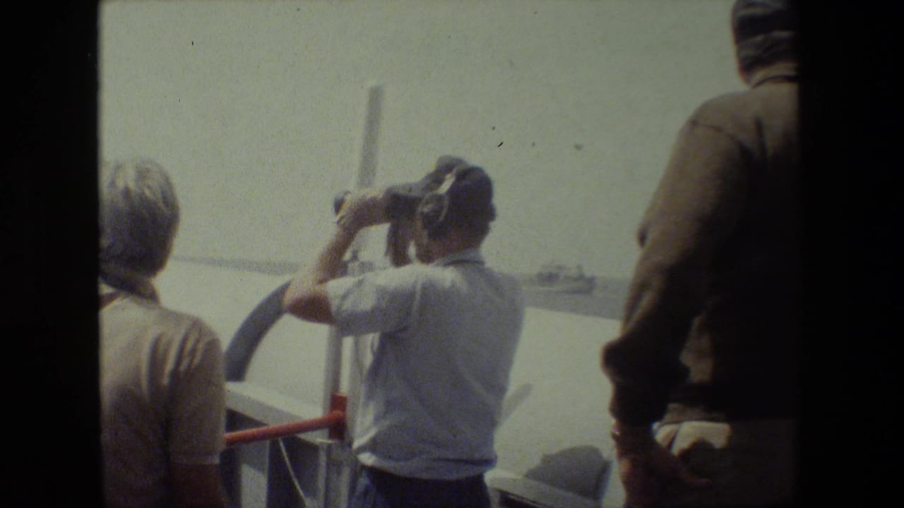 Adults on a ship deck watch the horizon while one man uses binoculars.