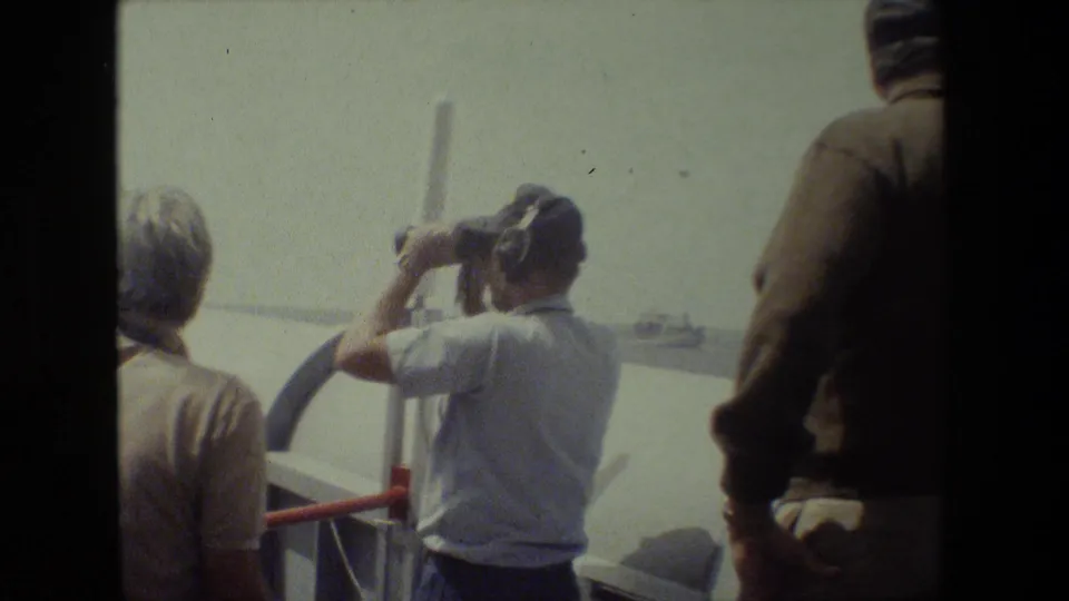 Adults stand near ship railings at sea as a man in a light shirt raises binoculars toward a distant vessel on the horizon.