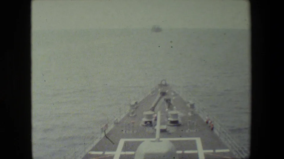 Forward-looking shipboard view over the bow and deck fittings toward open ocean, with a small vessel faintly visible near the horizon.