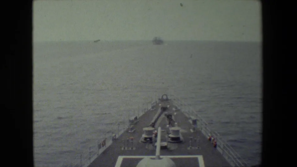 Faded 8mm frame looking down the bow of a moving ship across open water, with deck equipment and a distant vessel still visible ahead.