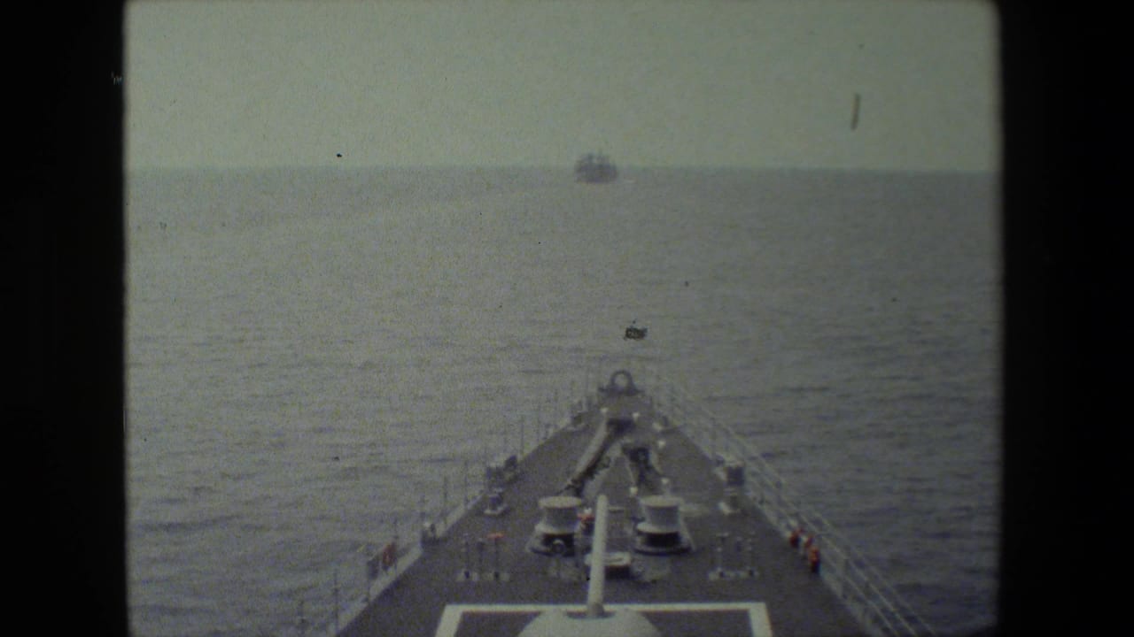 Forward shipboard view over a naval vessel's bow, with deck equipment, railings, open ocean, and small distant vessels under a hazy sky.