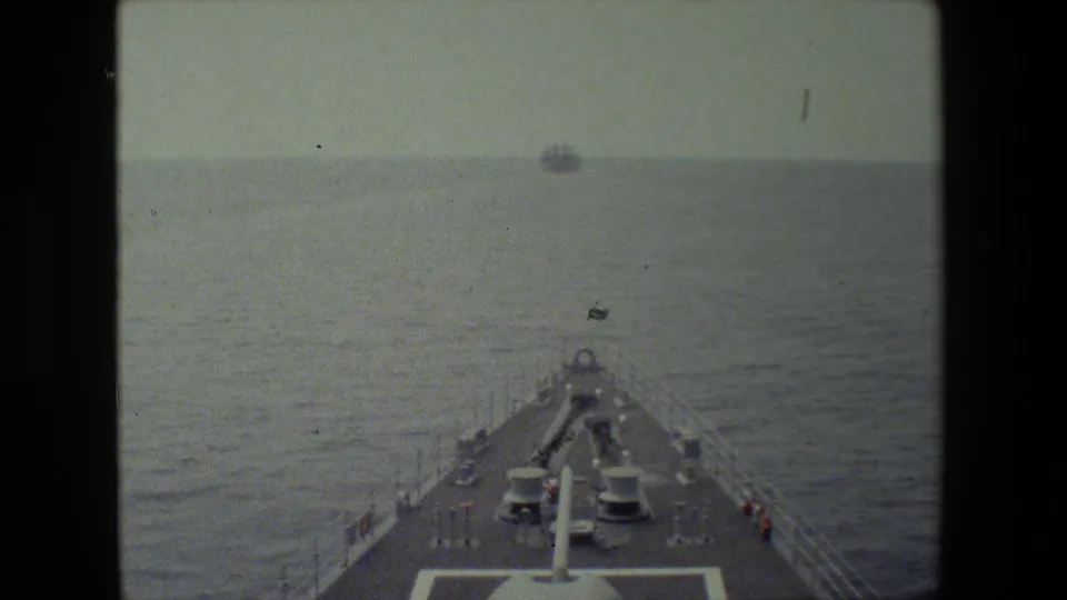 Forward shipboard view over a naval vessel's bow, with deck equipment, railings, open ocean, and small distant vessels under a hazy sky.