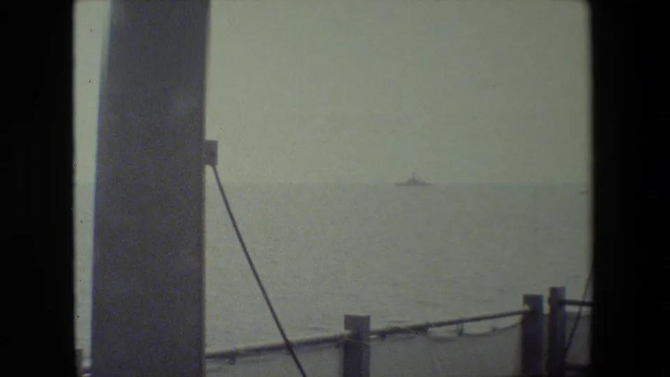 A hazy ocean horizon with a distant vessel, framed by vertical ship structure, rigging or cable, and deck railing in the foreground.