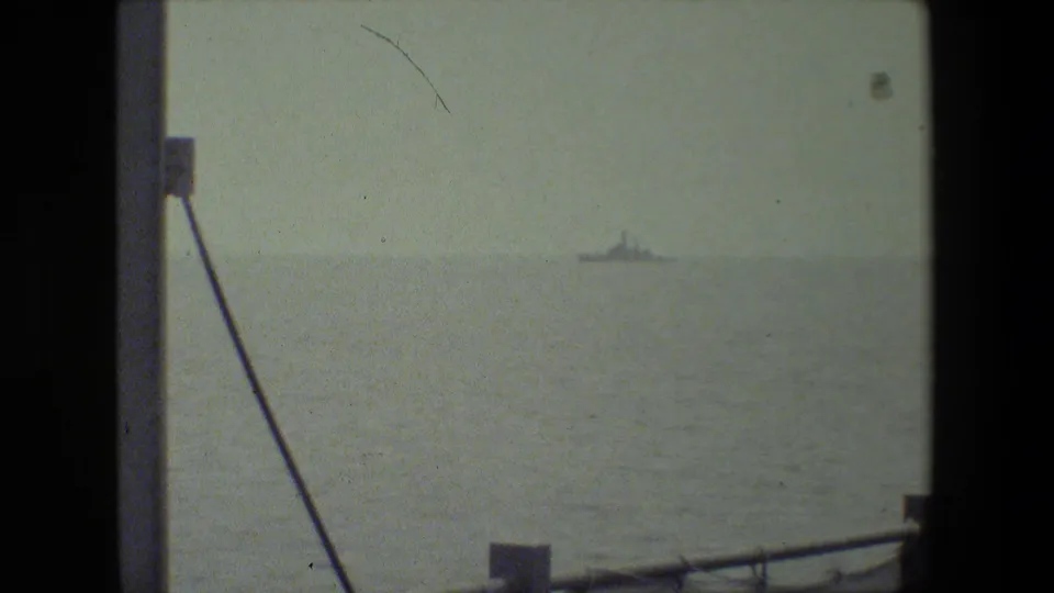 Shipboard view across pale water toward a distant vessel, with a diagonal cable, partial vertical structure, and low railing in the foreground.