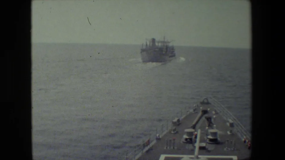 A larger vessel moves across open water ahead of a naval ship bow, with deck fittings and the hazy horizon visible in grainy 8mm footage.