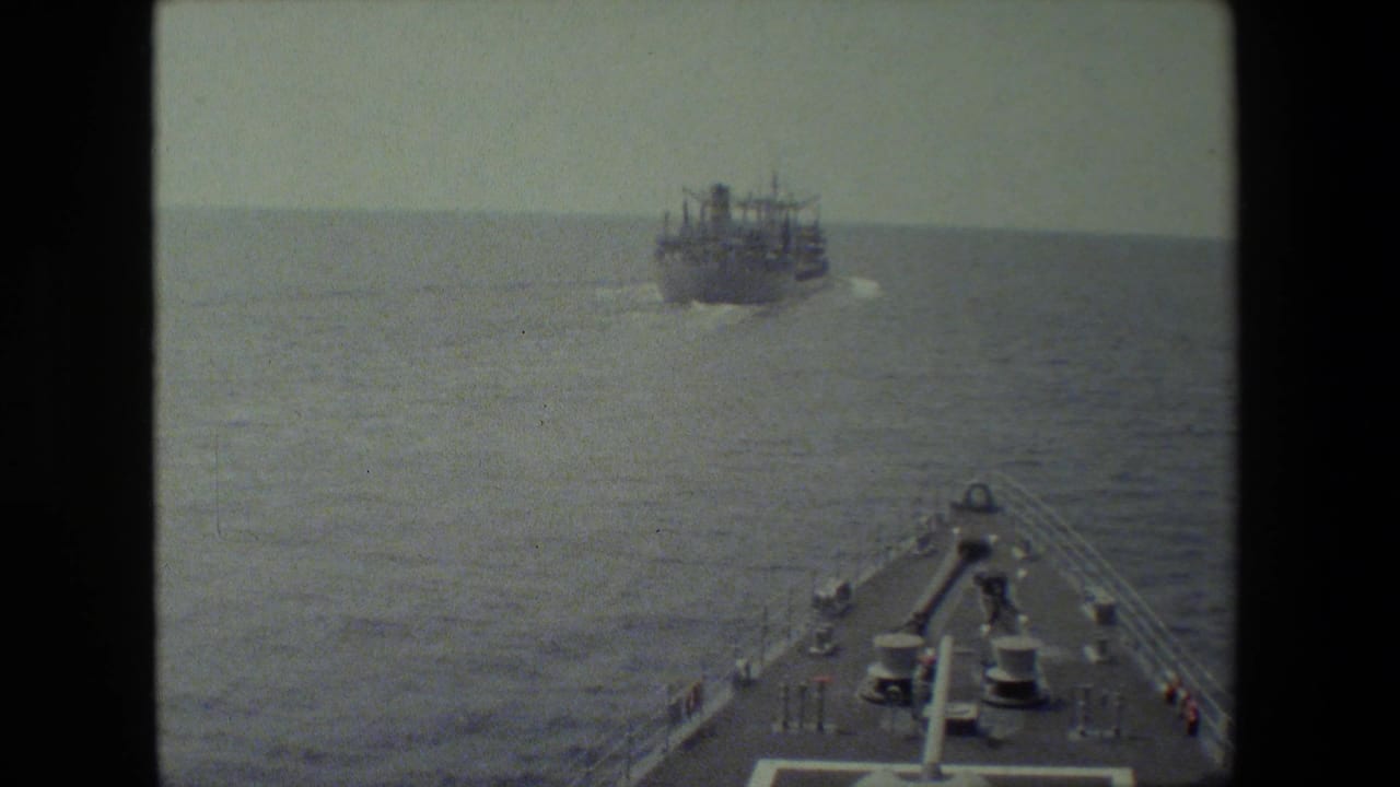 A forward deck view from a vessel at sea shows railings and deck equipment in the foreground with a large ship moving on the horizon.
