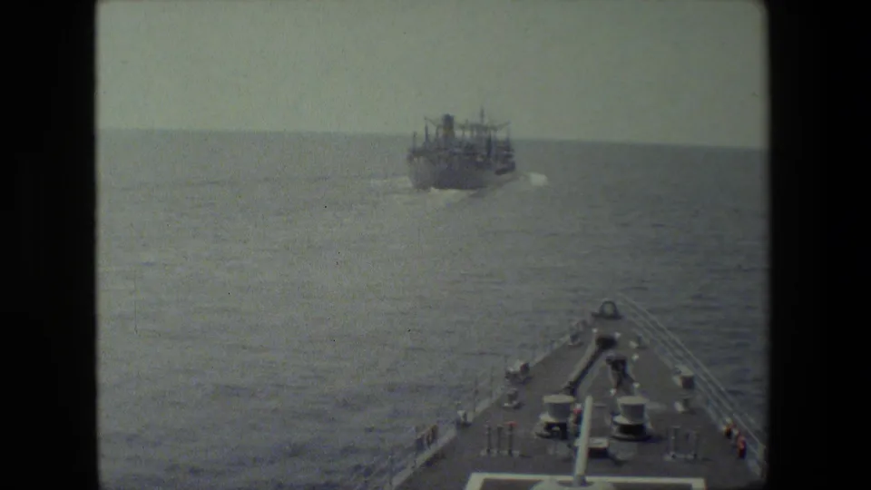 A forward deck view from a vessel at sea shows railings and deck equipment in the foreground with a large ship moving on the horizon.