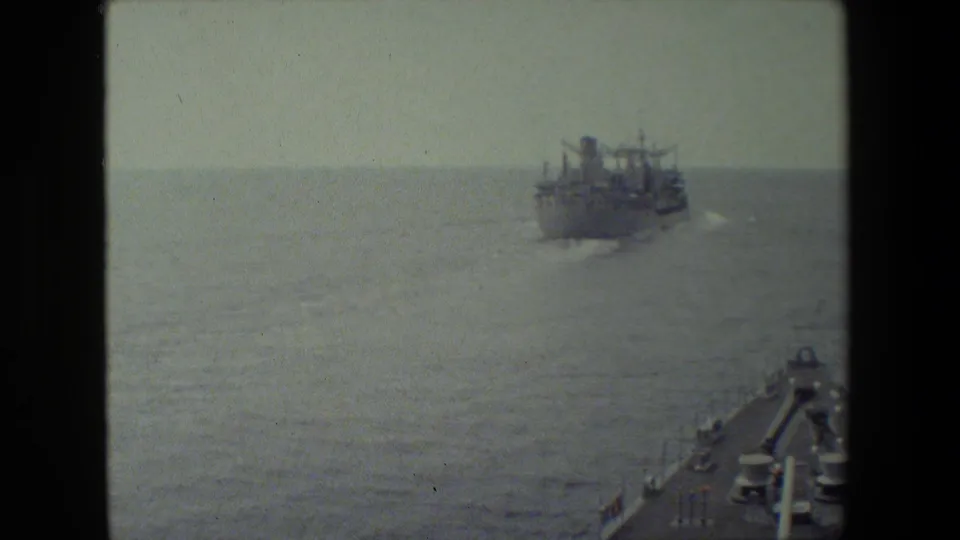 The bow area of the filming vessel remains at lower right while a large naval or supply ship travels across the water leaving a pale wake.