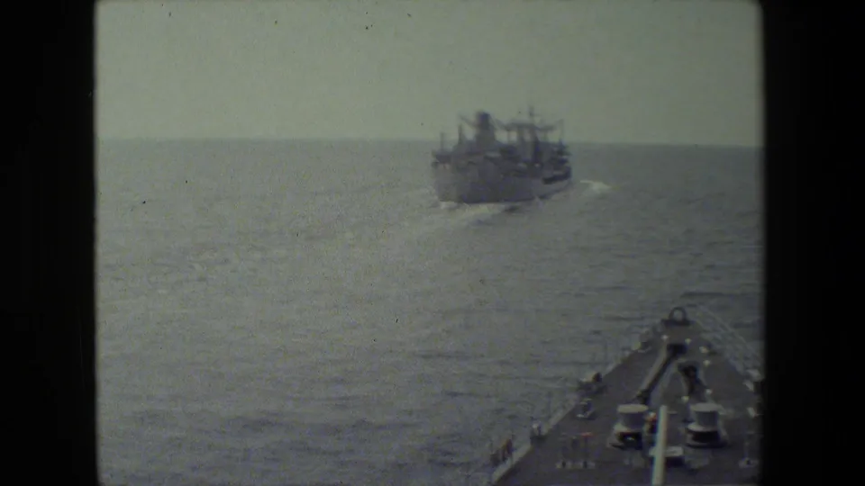 A handheld deck view frames the ocean and a distant large ship, with the filming vessel's forward structure visible at the lower right edge.