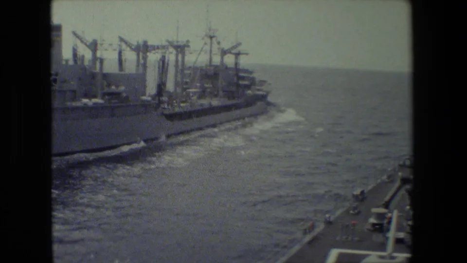 A closer view shows a large naval or supply ship alongside the filming vessel, with cranes or upright structures on deck and wake along the hull.