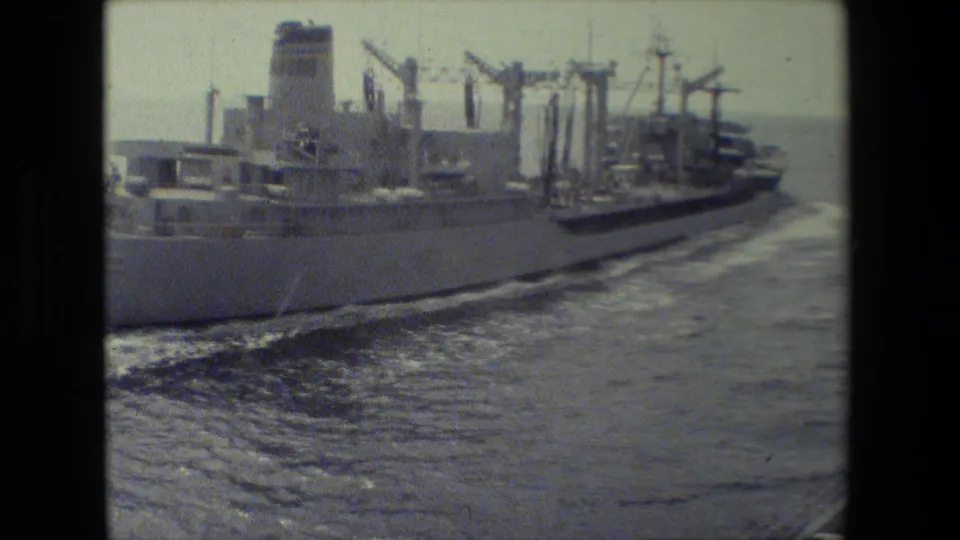 The ship's side hull, deck equipment, and wake are visible as it moves through open water in a grainy 8mm frame.