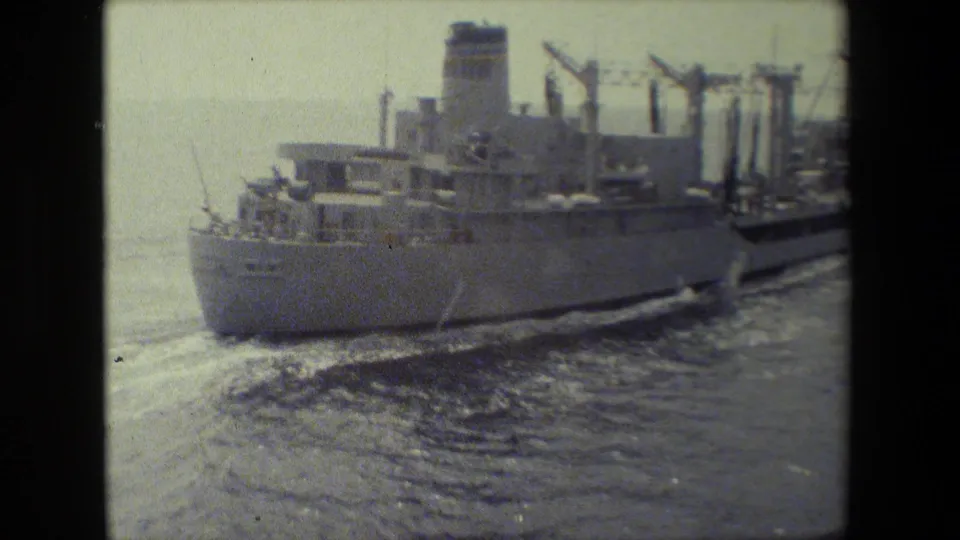 The stern and upper deck of the large ship pass through the frame, with cargo-handling structures and turbulent wake visible behind it.