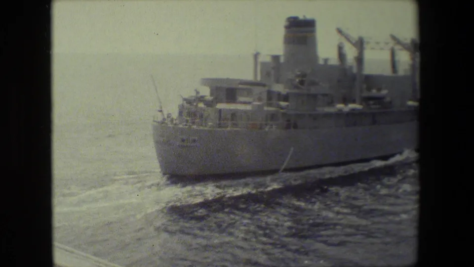 The rear side of the naval cargo or support ship recedes across bright open water, with its superstructure, railings, and foamy wake visible.