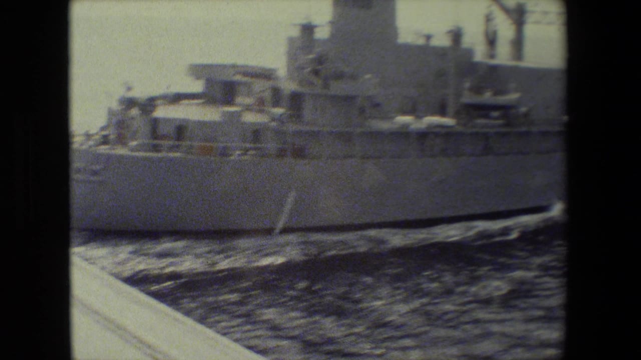 A large gray military ship fills the frame as it moves across open water, with the hull, superstructure, deck equipment, and wake visible in grainy 8mm footage.