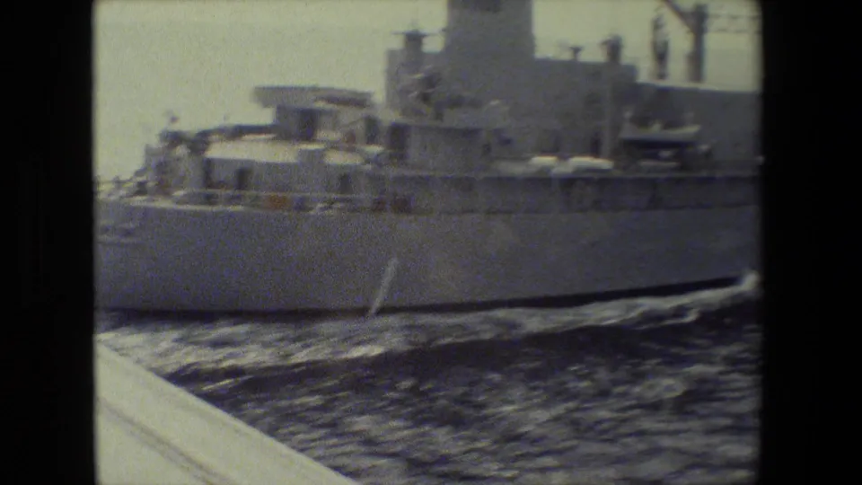 A large gray military ship fills the frame as it moves across open water, with the hull, superstructure, deck equipment, and wake visible in grainy 8mm footage.
