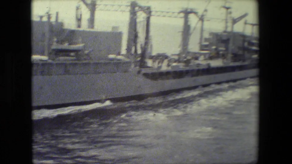 The rear portion of the gray military ship moves through the frame, with cranes, railings, small deck figures, and disturbed water visible in the 8mm scan.