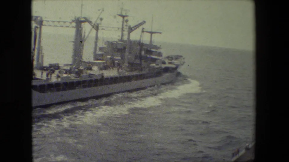 The ship is seen farther aft as it continues across open water, with cranes, mast structures, deck areas, and a broad foamy wake trailing behind.