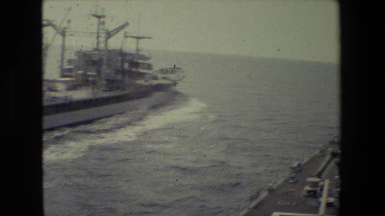 A moving-vessel view shows a large ship to the left and the edge of another ship at lower right as both travel across choppy open water in archival 8mm footage.