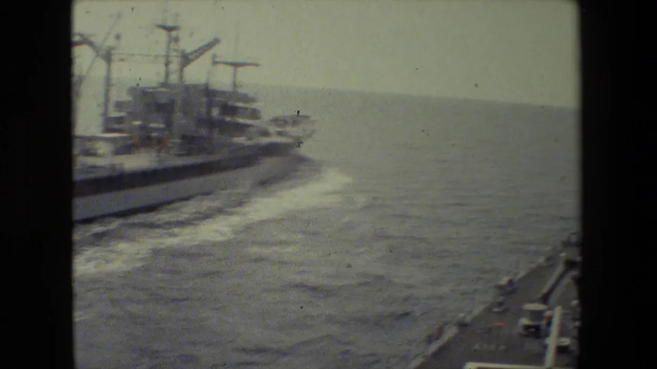 A moving-vessel view shows a large ship to the left and the edge of another ship at lower right as both travel across choppy open water in archival 8mm footage.