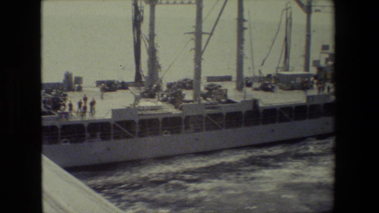 A military ship with cranes and deck equipment passes close beside the camera vessel.