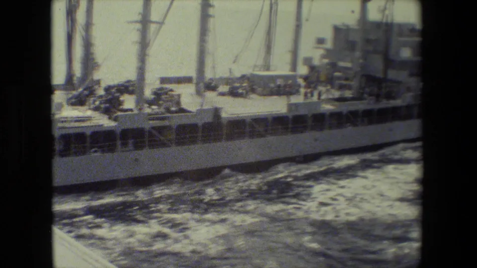 The camera holds a side view of the vessel's deck, showing cranes or masts, equipment, railings, distant crew figures, and a white wake along the hull.