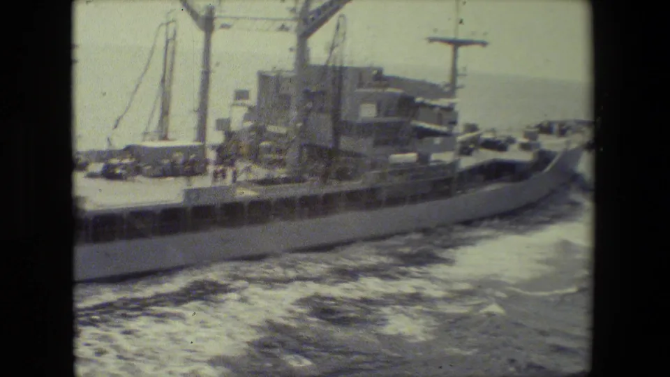 A military-looking vessel's stern area and deck structures pass through foamy water, with cranes, equipment, railings, and open sea visible beyond.