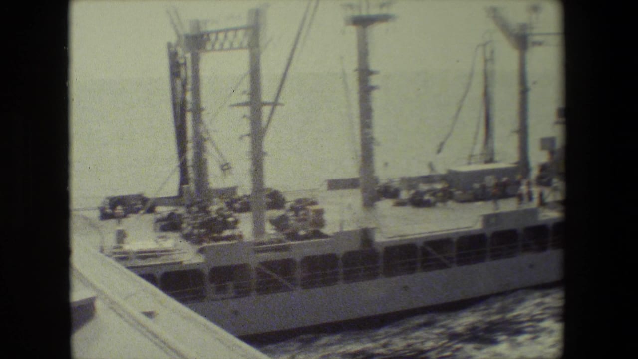 A large naval vessel passes near the camera, showing masts, deck equipment, railings, and wake beside the hull in soft archival film.