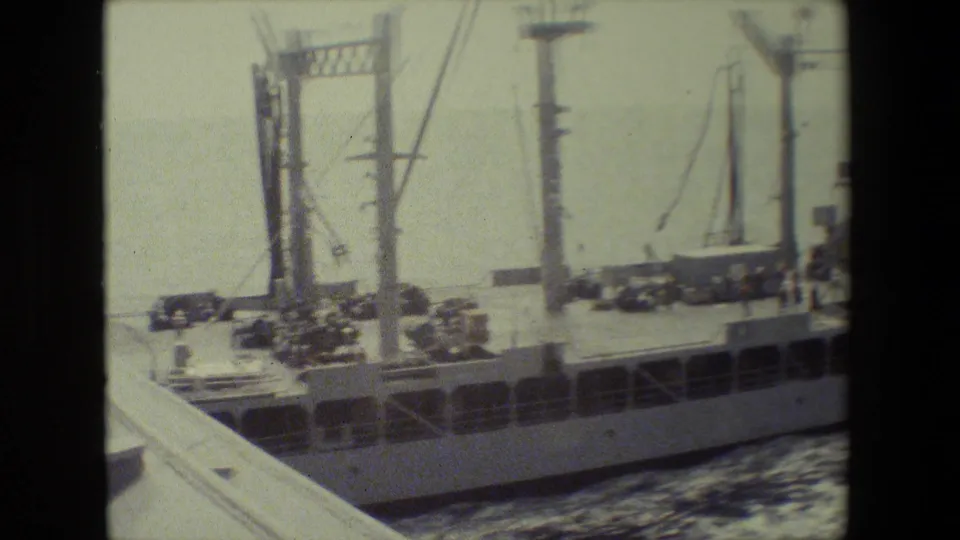A large naval vessel passes near the camera, showing masts, deck equipment, railings, and wake beside the hull in soft archival film.