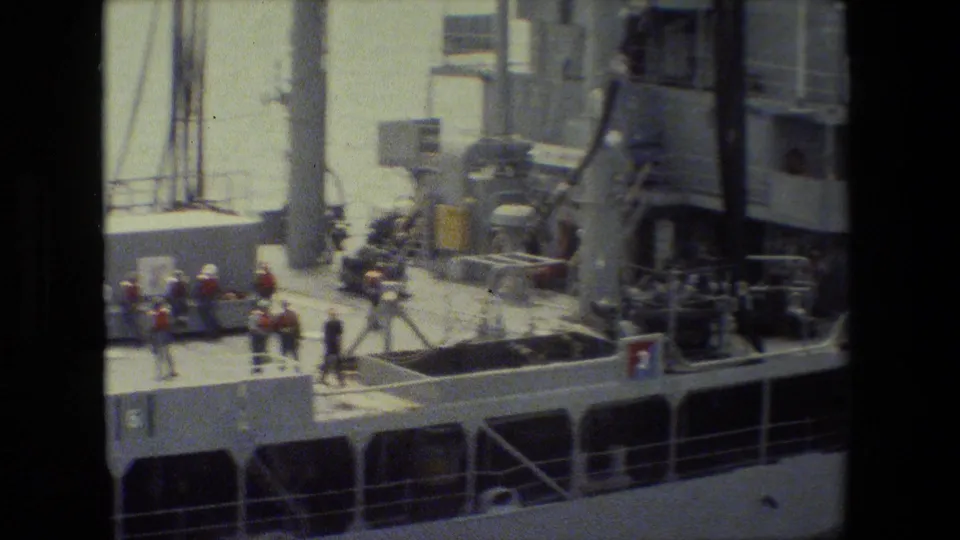 Crew members in light and darker clothing stand and move on the deck near ship equipment, railings, masts, and the vessel superstructure.