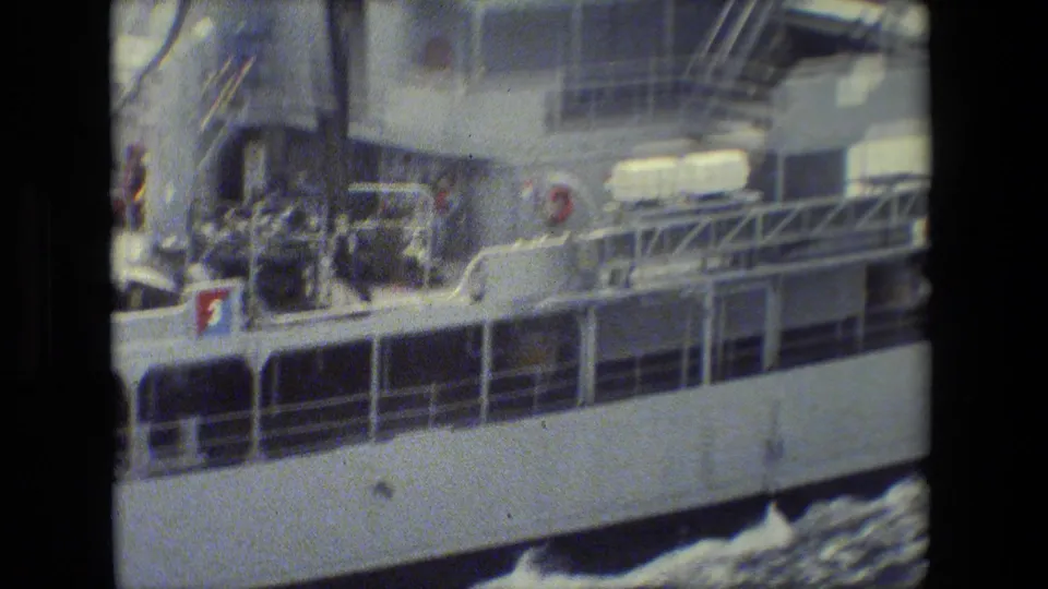 The side of the large naval vessel fills the frame, with crew visible near railings and deck structures above water churned along the hull.