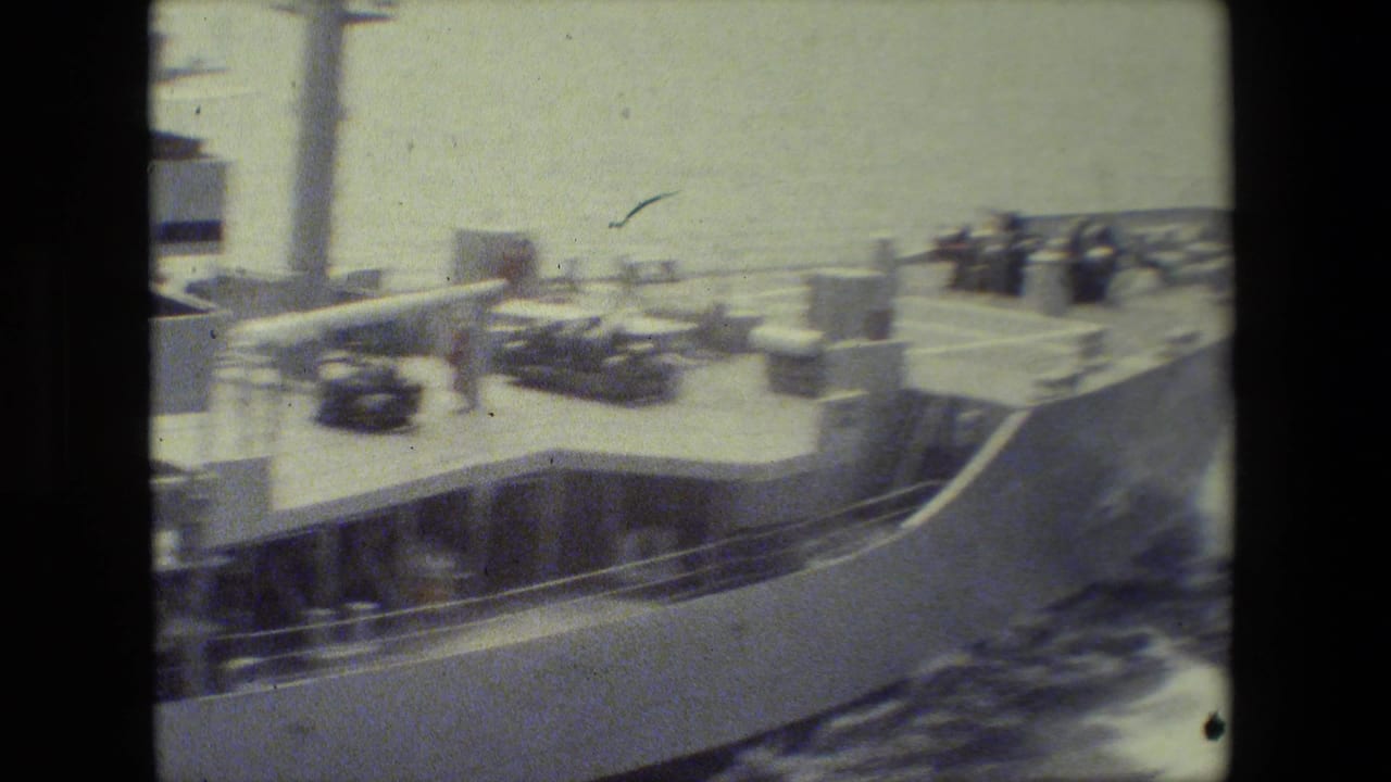 Faded 8mm frame of a vessel deck with railings, small equipment or vehicles, a few distant people, and water spray along the side.