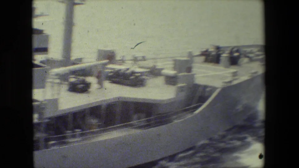 Faded 8mm frame of a vessel deck with railings, small equipment or vehicles, a few distant people, and water spray along the side.