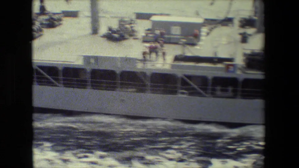 Blurred 8mm frame showing the vessel's side, rectangular openings or windows, deck gear, distant figures, and dark water moving below the hull.