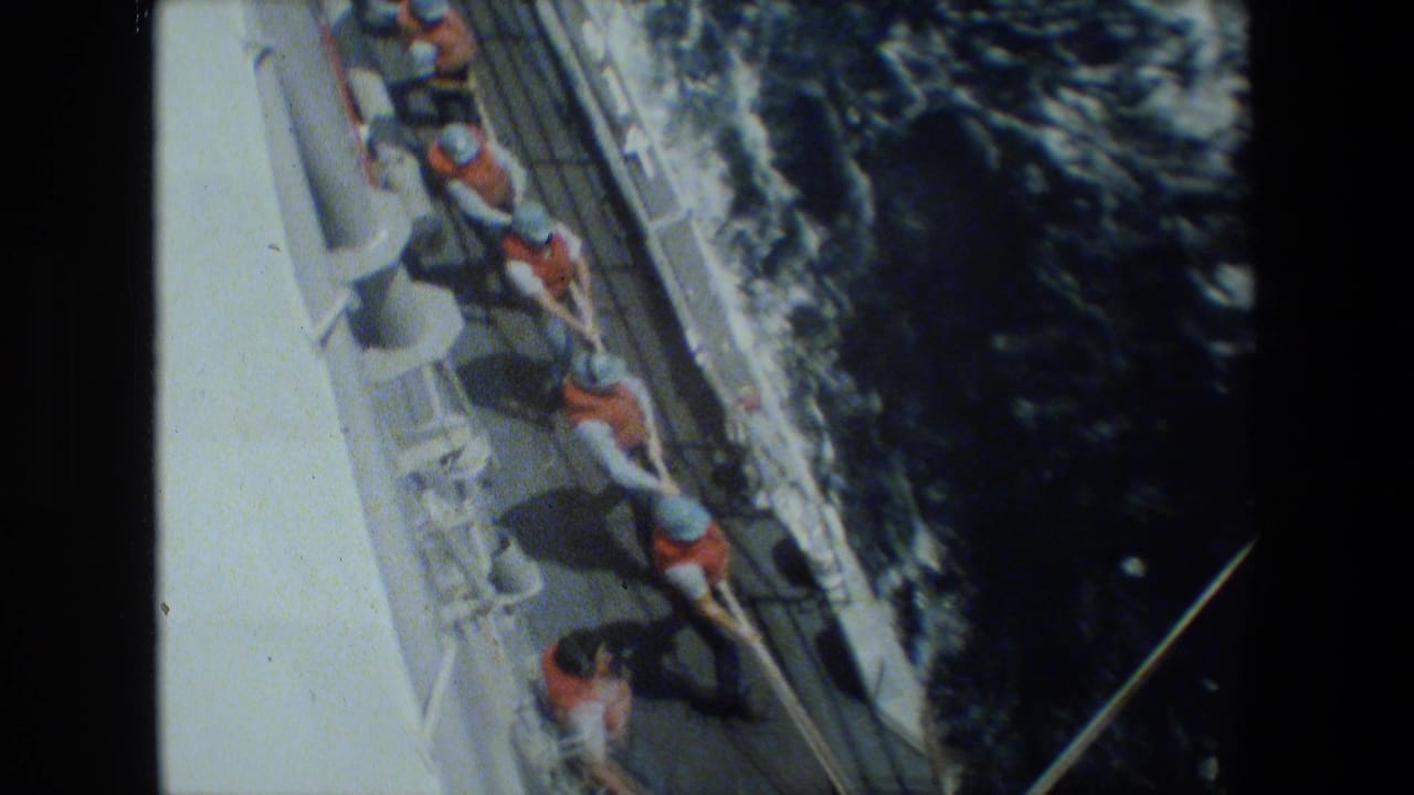 High-angle 8mm frame of sailors in orange life vests and light hats pulling a thick line along a ship deck, with dark water and wake beside the vessel.