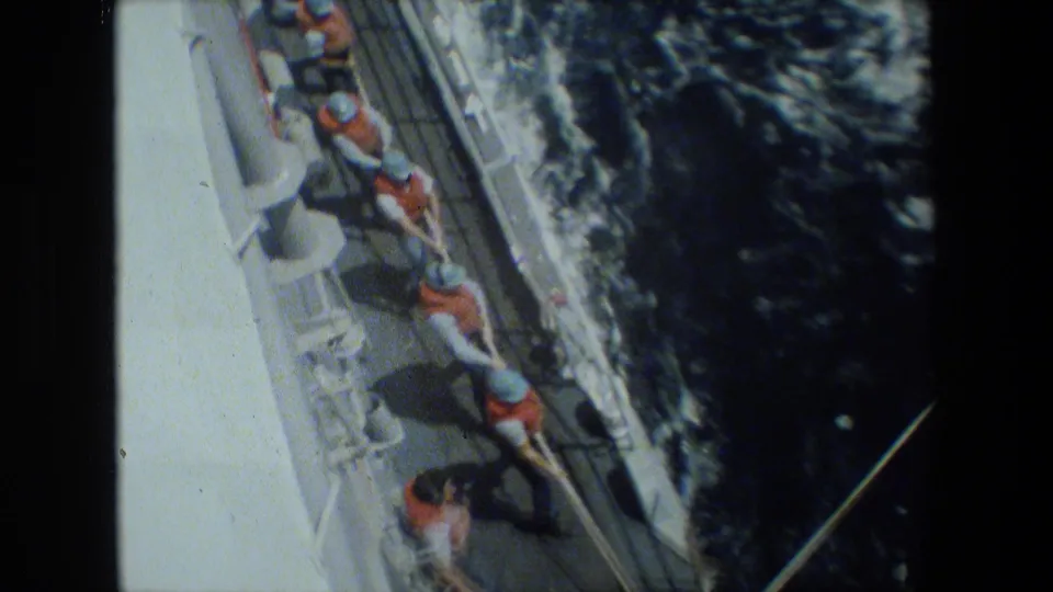 High-angle 8mm frame of sailors in orange life vests and light hats pulling a thick line along a ship deck, with dark water and wake beside the vessel.